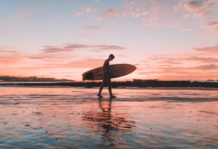 junger Mann trägt ein Surfbrett am Strand bei Sonnenuntergang